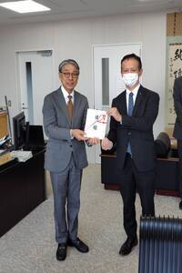 室内でスーツ姿の男性2人が並んで立ち、白い封筒を、2人で一緒に手に持って正面を向いている写真