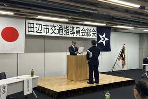 会場にて、前方の台にスーツ姿の男性が立ち、代表者から賞状を授与されている写真