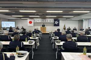 前方にスーツ姿の男性が立っており、並べられた机に座って多くの出席者が話を聞いている写真