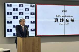 会見場にて挨拶と名前が映し出されたモニターがある中で、スーツ姿の男性がマイクでスピーチをしている写真