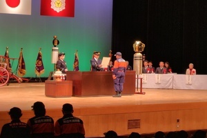 スーツ姿の男性が前方で、左手にマイクを持って前を見据えながら話している写真