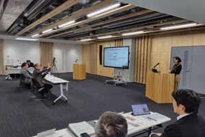 会場の右側で女性がマイクの前に立っており、前方に座っているスーツの男性が右手を挙げている写真